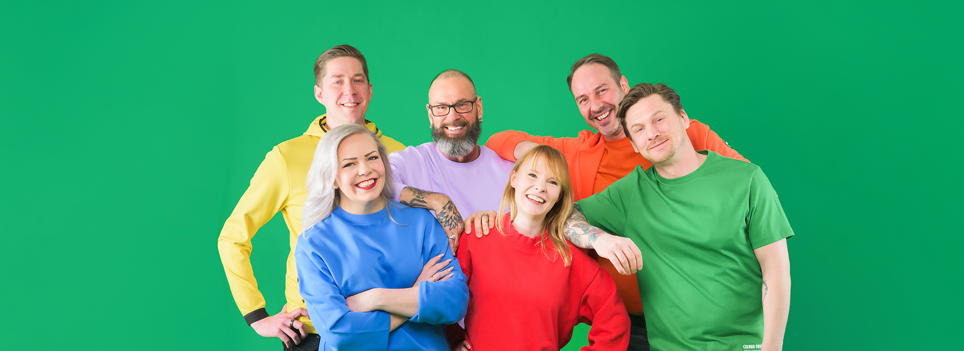 Kuusi värikkäissä vaatteissa olevaa henkilöä seisoo rykelmässä ja hymyilee kameralle.